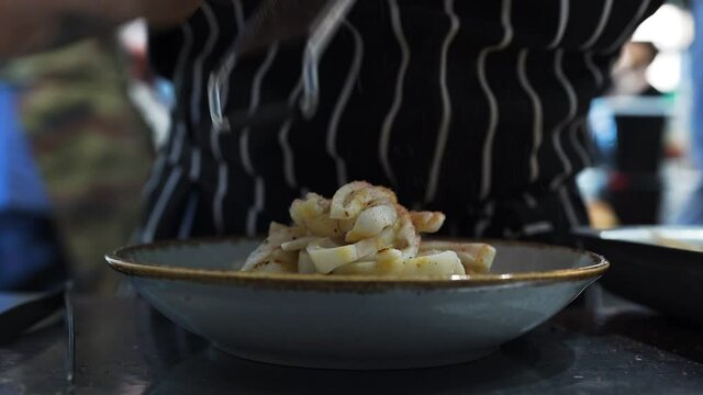 Bustle of restaurant kitchen weekends. Fried squid ring, chef finely grates basturma onto dish. Working atmosphere, recipes from menu are prepared inside the restaurant business. Social media content.