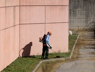 A gardener trims the grass on a  sidewalk with a weed eater. He wears all the prescribed safety clothes and accessories.