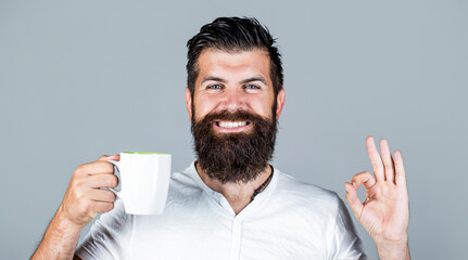 Handsome man holds cup of coffee, tea. Bearded man smiling showing sign ok. Good morning, man tea, ok. Smiling hipster man with cup of fresh coffee, Happy man showing ok sign. Morning concept