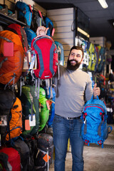 Smiling man customer examining backpacks in sports equipment store.