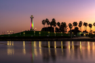 Sunset over illuminated Long Beach Lighthouse, palm trees and grassy area at ShoreLine Aquatic Park, with shimmering reflections in the water.