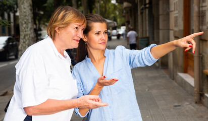 Polite young woman pointing way to middle aged female tourist on summer city street..