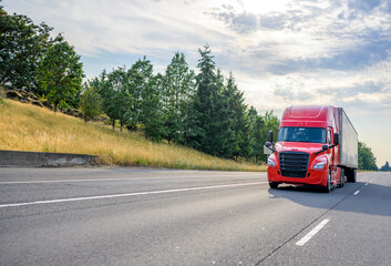 Red big rig semi truck with black grille transporting cargo in semi trailer driving on the straight wide highway road with trees on the hillside