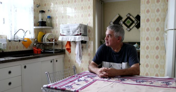 Thoughtful older man sitting at kitchen table. Pensive middle aged senior person