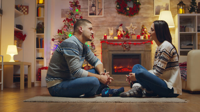 Beautiful young couple looking at each other sitting on the carpet celebrating christmas in front of fireplace.