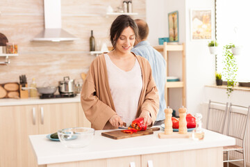 Obraz na płótnie Canvas Girlfriend slicing red bell pepper for salad in kitchen. Boyfriend in the background. Beautiful cheerful happy smiling woman in bright room preparing healthy and organic meal.