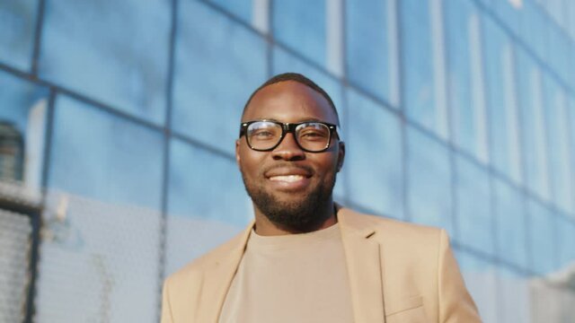 Chest-up shot of joyous African American businessman in smart casual wear and glasses looking at camera, smiling and laughing while standing in front of glass building in city on sunny summer day