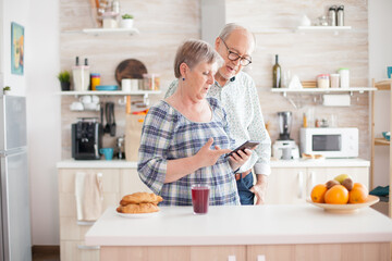 Obraz na płótnie Canvas Caucasian senior wife and husband in kitchen during breakfast using smartphone technology and internet conection. Leisure retired woman and man smiling in domicile, relaxing.