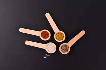 Salt pepper chilli curry oregano powders in wooden spoons on black stone background