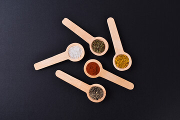 Salt pepper chilli curry oregano powders in wooden spoons on black stone background
