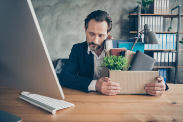 Photo of dismissed sad worker mature guy fired agent jobless loser eyes closed packed belongings hold big carton box exhausted financial crisis crash sit workstation modern office indoors