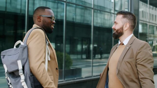 Cheerful Caucasian businessman walking outdoors in city downtown, meeting African American male colleague, greeting him with handshake and chatting