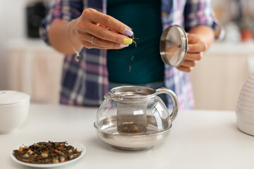 Close up of pouring aromatic herbs in teapot to make tea in morning for breakfast. Preparing tea in...