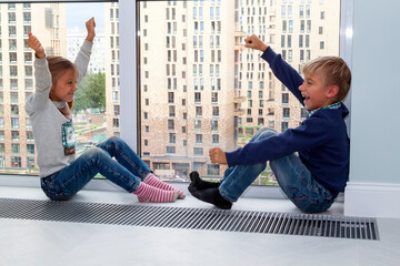 two children with their hands in air rejoice at end of quarantine. girl with pigtails and boy look at each other happily sitting on floor by panoramic window