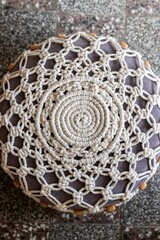 Pouf covered with a cover made in macrame technique from white cotton cord with wooden beads reflected top view on the background of a stone floor, boho style