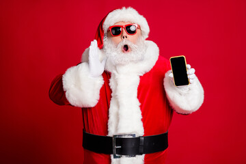 Photo of pensioner old man grey beard hold telephone empty space hand open mouth yell sale information wear x-mas santa costume gloves coat belt sunglass cap isolated red color background