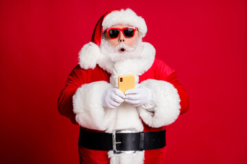 Photo of retired old man grey beard hold telephone shocked impressive news north pole flight available wear santa x-mas costume gloves coat sunglass headwear isolated red color background