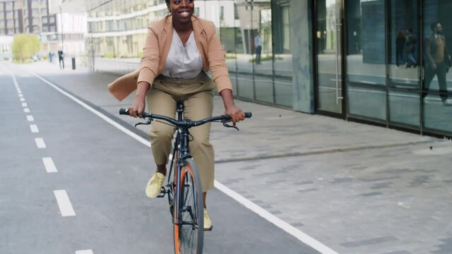 Young happy Afro-American businesswoman riding bicycle along urban street in the city and smiling while going to work in the morning