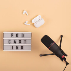 Podcasting lettering next to microphone and wireless headphones at workplace on beige background.