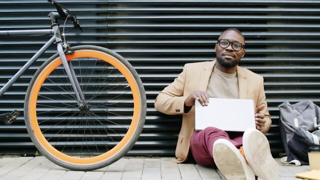 Medium shot of young Afro-American businessman sitting on ground beside bike, browsing the Internet, then closing laptop and looking at camera with smile while working outdoors on street