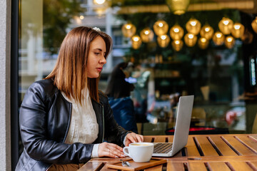Fototapeta premium Outdoor portrait of elegant caucasian female sitting at modern restaurant, working on her laptop and drinking coffee. Looking at computer. Romantic woman blogger. Freelancer thinking about new ideas.