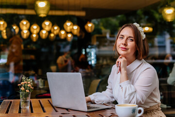 Attractive young elegant caucasian business woman works freelance on laptop computer, sits alone at cafe. Cafe culture, casual lifestyle. Romantic female blogger. Freelancer thinking about new ideas.