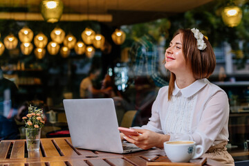 Attractive young elegant caucasian business woman works freelance on laptop computer, sits alone at cafe. Looking away smiling. Student learning online