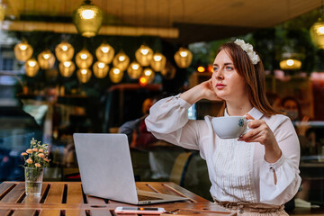Attractive young elegant caucasian business woman sits alone at cafe, holding coffee cup. Looking away smiling. Student learning online. Romantic female blogger. Freelancer thinking about new ideas.