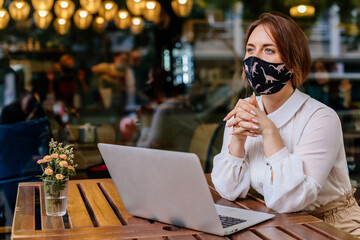 Casual caucasian business woman wearing protective mask working on laptop in a cafe. COVID – 19 virus protection at work. The concept of remote work and online distance learning