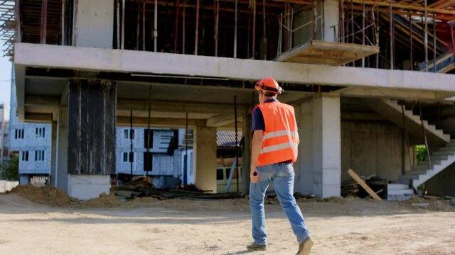 At big construction site walking foreman in a safety equipment to the construction site taking video from the back