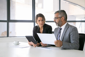 Company leaders analyzing reports. Two business colleagues sitting together, looking at document, holding tablet and talking. Medium shot. Communication concept
