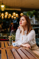 Young beautiful woman holding a cup of coffee in cafe. Outdoor portrait of female blogger posing while traveling holiday vacation. Happy freelancer thinking about new ideas.  Looking away