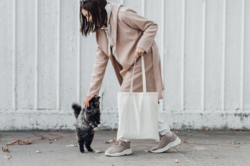 Woman in coat with white cotton bag in her hands and cute cat sitting near. Mockup and zero waste concept. - obrazy, fototapety, plakaty