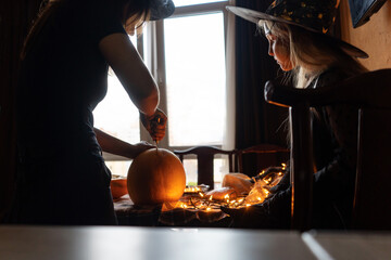 Woman and child girl having fun and celebrate Halloween in witch costume. Mother and daughter carving pumpkin. Back light.