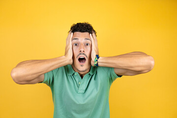 Young handsome man wearing green casual t-shirt over isolated yellow background crazy and scared with hands on head, afraid and surprised of shock