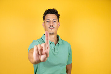 Young handsome man wearing green casual t-shirt over isolated yellow background showing and pointing up with fingers number one while is serious