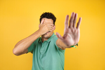 Young handsome man wearing green casual t-shirt over isolated yellow background covering eyes with hands and doing stop gesture with sad and fear expression. Embarrassed and negative concept.