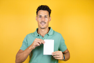 Young handsome man wearing green casual t-shirt over isolated yellow background smiling and showing blank notebook in her hand