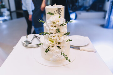 A beautiful white three-tiered cake decorated with flowers and a dish for dessert with a knife stands on a table in a restaurant at a wedding.