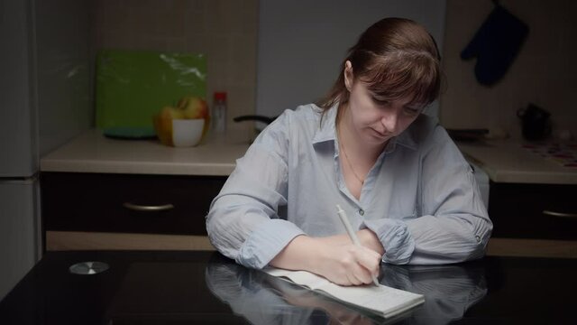 serious woman is sitting in the kitchen in the evening thinking and writing in a notebook