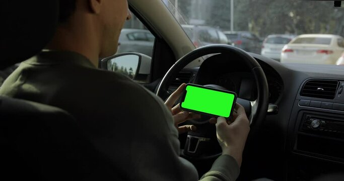 Over the shoulder view. Man in a sweater sitting in the car and watching iPhone 11 with green screen. Landscape horizontal orientation. Application promo. Fall background.