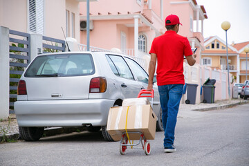 Postman holding tablet and wheeling trolley with carton boxes. Caucasian professional courier in red uniform walking on street with cardboard parcels on cart. Delivery service and post concept