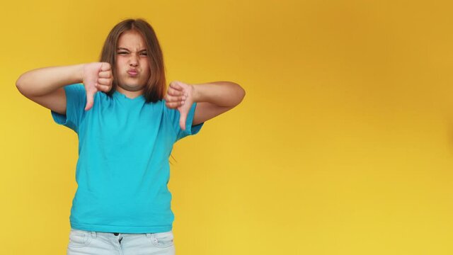 Dislike gesture. Disgusted kid. Wrong choice. Bad idea. Skeptic disappointed young girl in blue t-shirt denying offer with thumbs down isolated on yellow copy space background.
