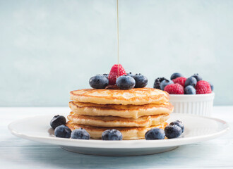 Stack of pancakes with fresh blueberries served in a dish with maple syrup being poured. Delicious breakfast.