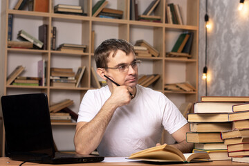 a young man in glasses sits at a table with a laptop, books and papers. office employee at work looks away with a thoughtful look. European appearance.