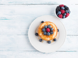 Stack of pancakes with fresh blueberries served in a dish with maple syrup. Delicious breakfast top view on white wooden table.