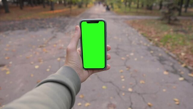Man hand holding iPhone in vertical or portrait orientation. People walking in the fall / autumn park. Pre-Keyed green screen ready for your content. POV, point of view. 