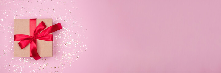 Gift box with red bow on festive pink background with golden sparkles around and copyspace for your text. Flat lay style. Christmas, New Year, Valentines Day or birthday celebration concept. Banner