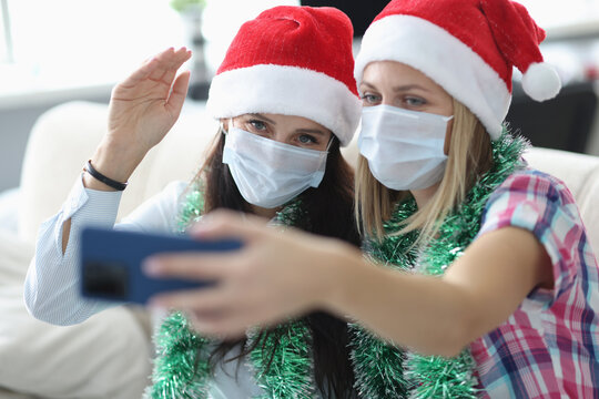 Two women in protective mask and santa claus hat talk online on phone. Girlfriend hold phone in her hand. Sister wave hello to screen.