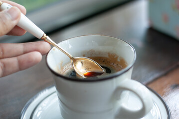 A hand holding siver spoon scooping some hot black coffee from a white ceramic mug